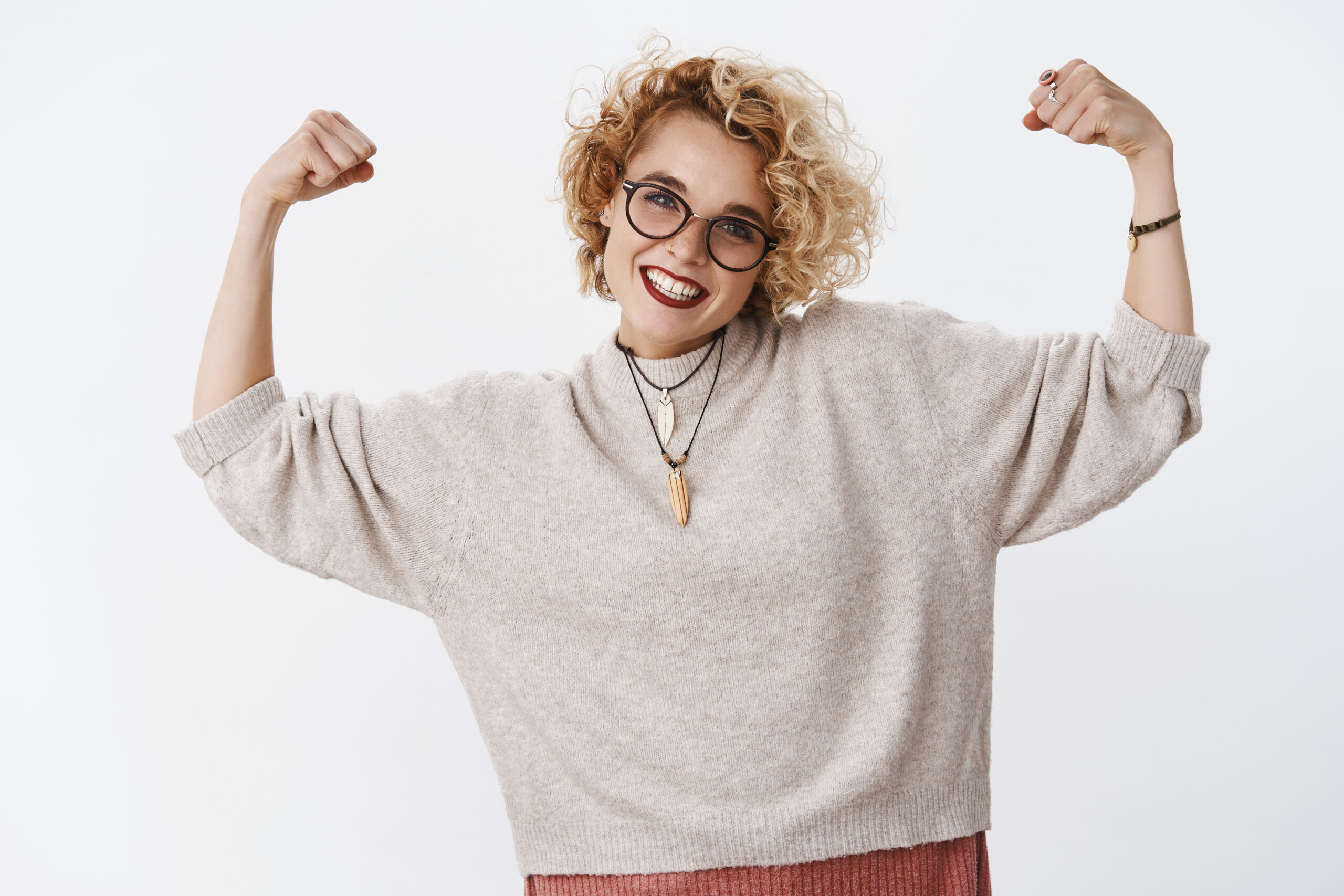 How strong girl is. Portrait of enthusiastic happy and confident european blond woman in glasses and sweater showing muscles and smiling joyfully showing power and courage over white background